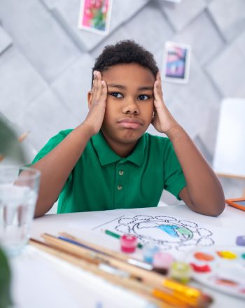 Meditation. Pensive dark-skinned boy of primary school age in green tshirt touching head with hands looking aside sitting at table with drawing and brushes in daylight